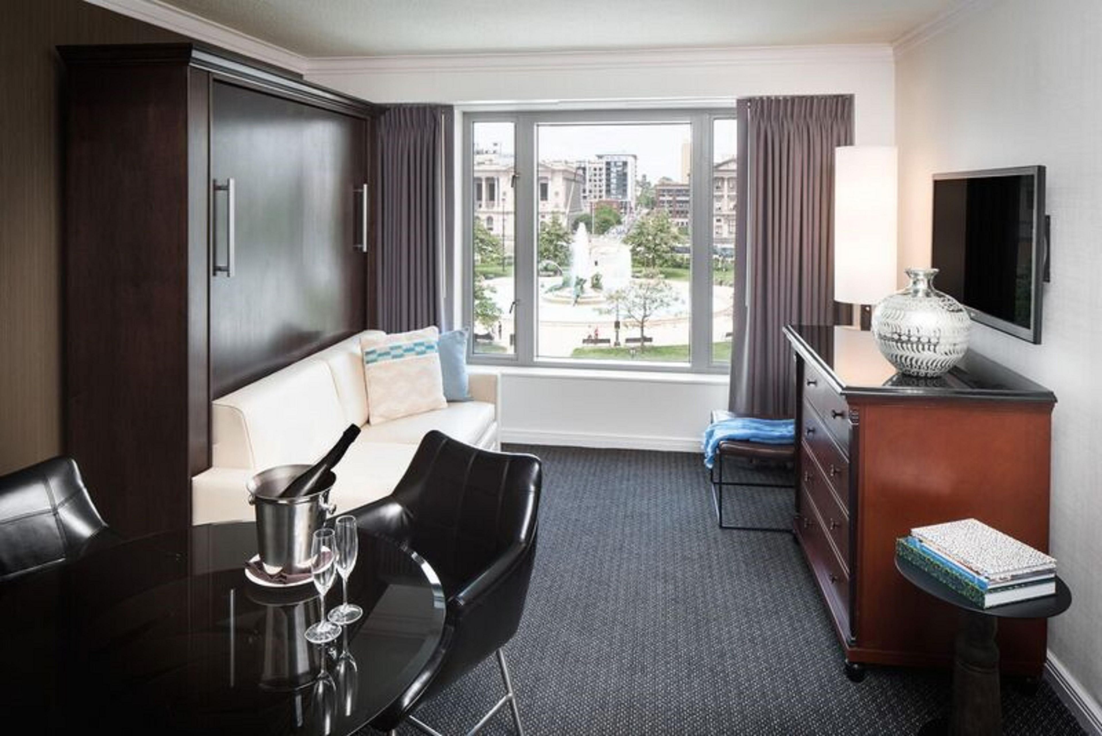 A hotel room with a sofa, a dining table with chairs, a TV, a lamp, and a view of the cityscape.