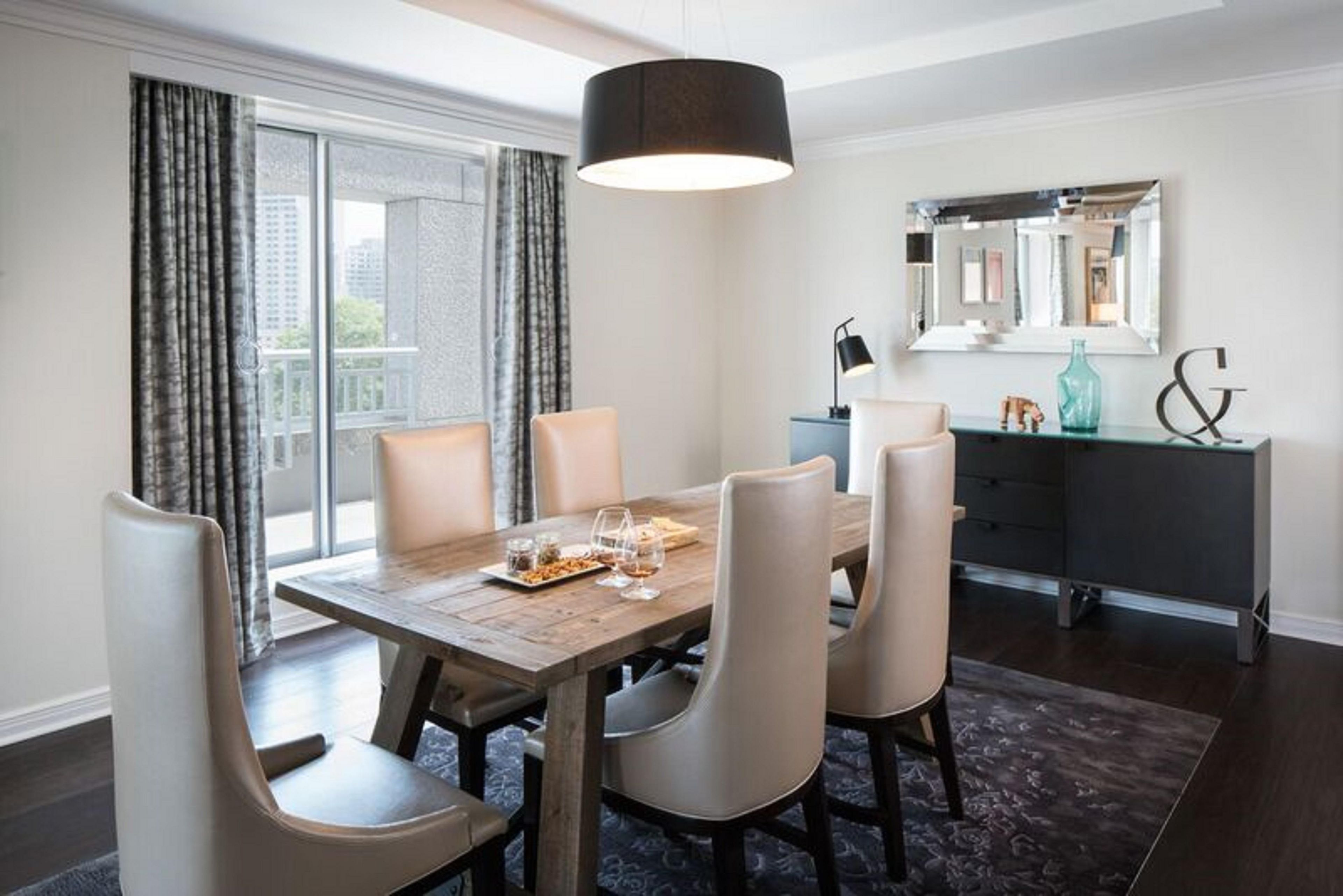Un coin repas avec une table en bois, six chaises, un luminaire suspendu noir et une vue dégagée grâce à de grandes fenêtres.