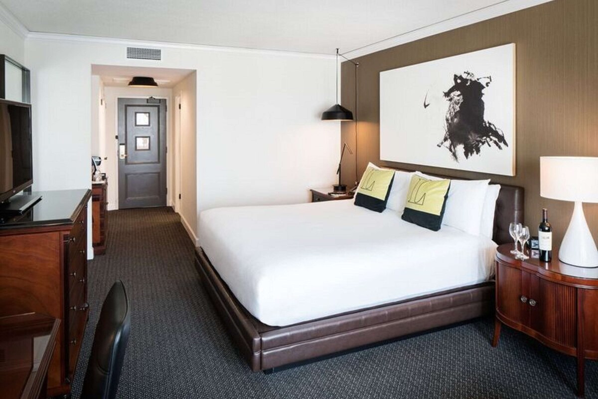 A hotel room with a large bed, a desk, a chair, a TV, a lamp, and a painting on the wall.