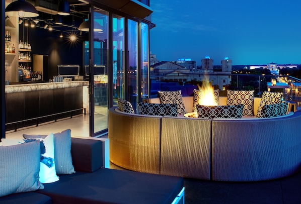 A rooftop bar with a fire feature, seating area, and city views.