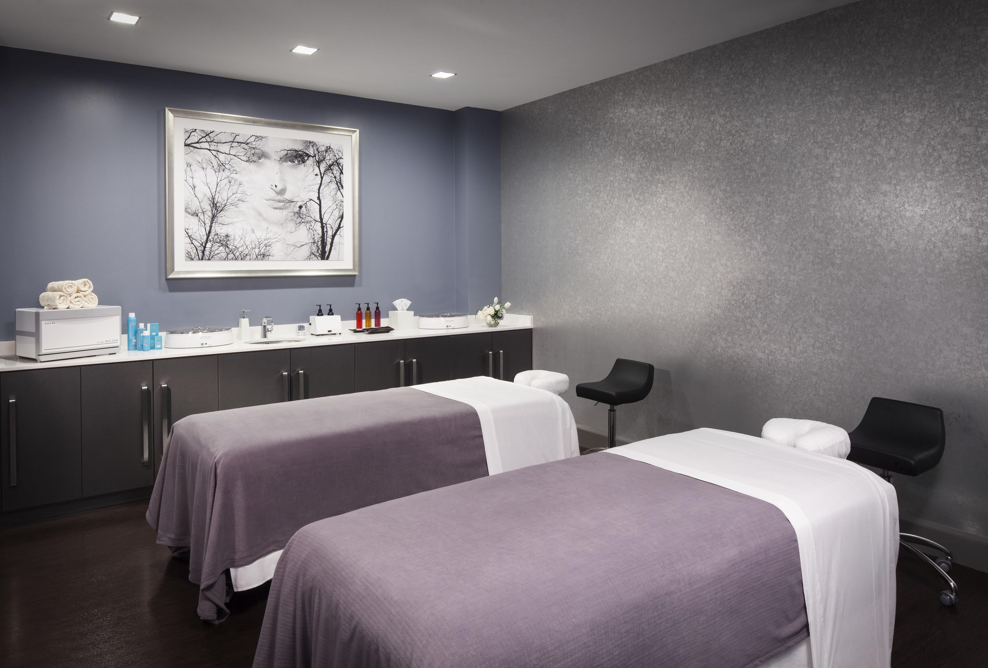 A modern spa room with two beds, a reception desk, and a large framed artwork on the wall.