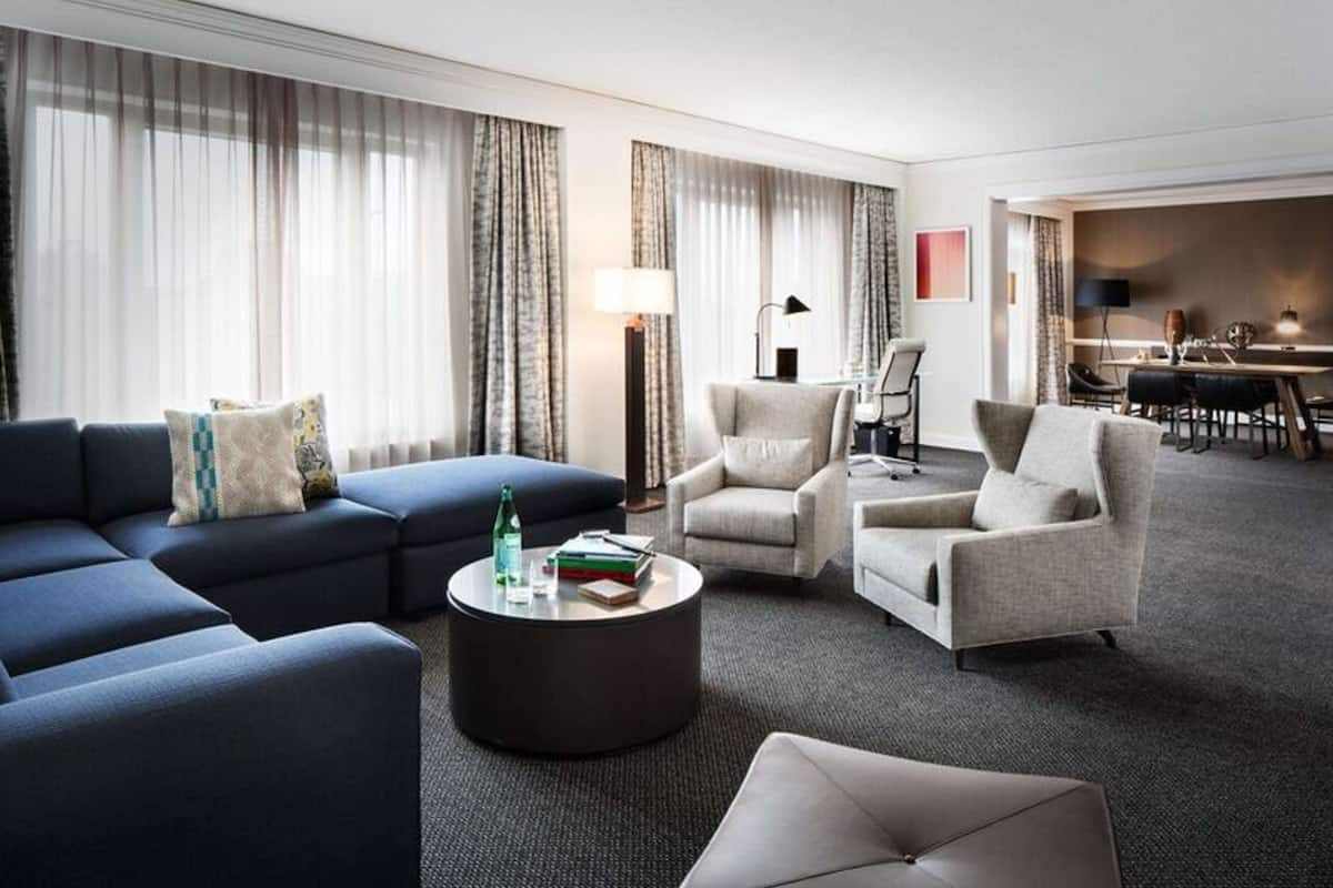 A modern hotel room with a blue sectional sofa, two armchairs, a round coffee table, and a dining area with a table and chairs.