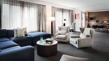 A modern hotel room with a blue sectional sofa, two armchairs, a round coffee table, and a dining area with a table and chairs.