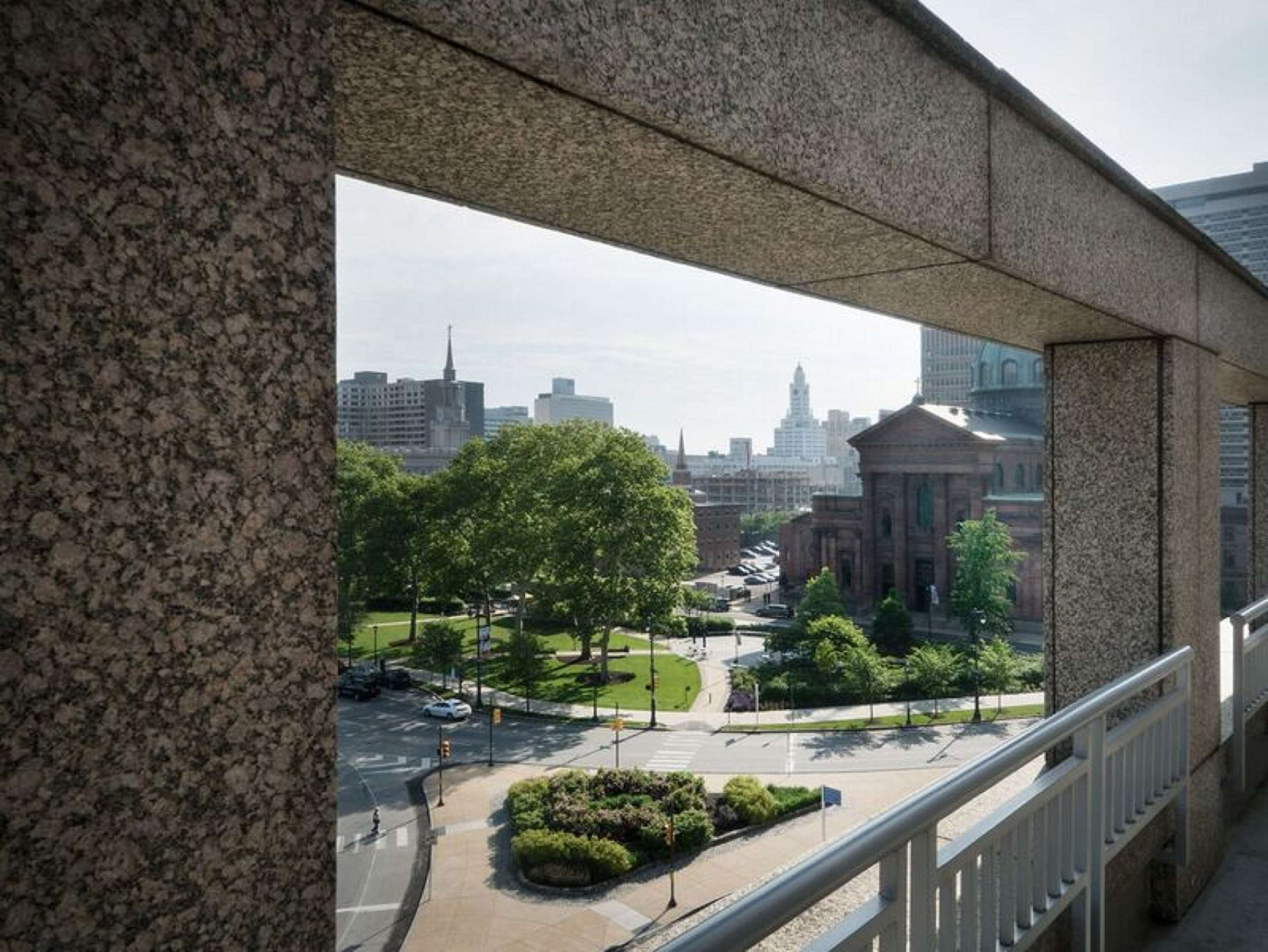 A view of a cityscape through a stone archway, including a large building with columns, a green park, and a crosswalk.