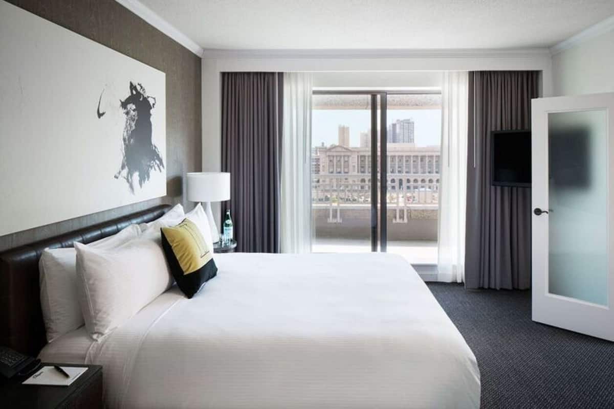 A hotel room with a large bed, a flat-screen TV, a painting on the wall, and a view of the city through the window.