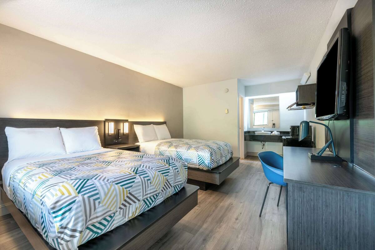 A hotel room with two beds, a desk, a chair, a TV, and a bathroom visible in the background.