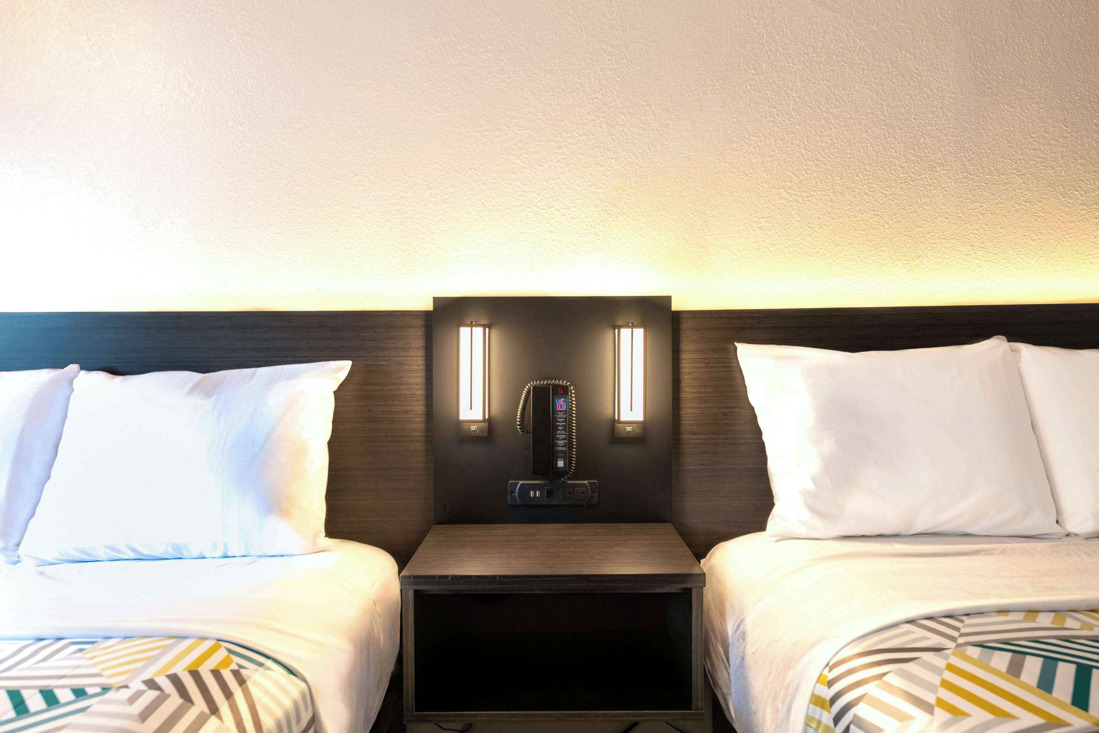 Une chambre d’hôtel avec deux lits, une table de chevet avec un téléphone et une horloge numérique, et deux lampes fixées au mur.