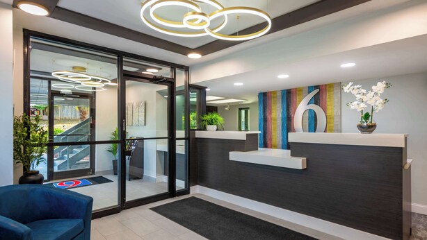 A modern reception area with a glass door entrance, a blue seating area, and a reception desk with a decorative number '6' sign. - Motel 6 Charlotte, NC - Coliseum (Charlotte)