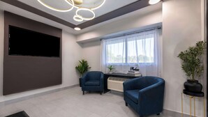 A modern living room with a flat-screen TV, two blue armchairs, a small table, a potted plant, and a window with sheer curtains. - Motel 6 Charlotte, NC - Coliseum (Charlotte)
