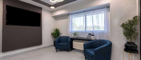 A modern living room with a flat-screen TV, two blue armchairs, a small table, a potted plant, and a window with sheer curtains.