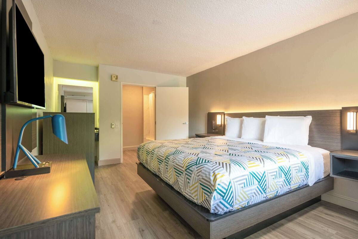 A modern hotel room with a large bed, bedside tables, a desk with a lamp, and a view of the hallway.