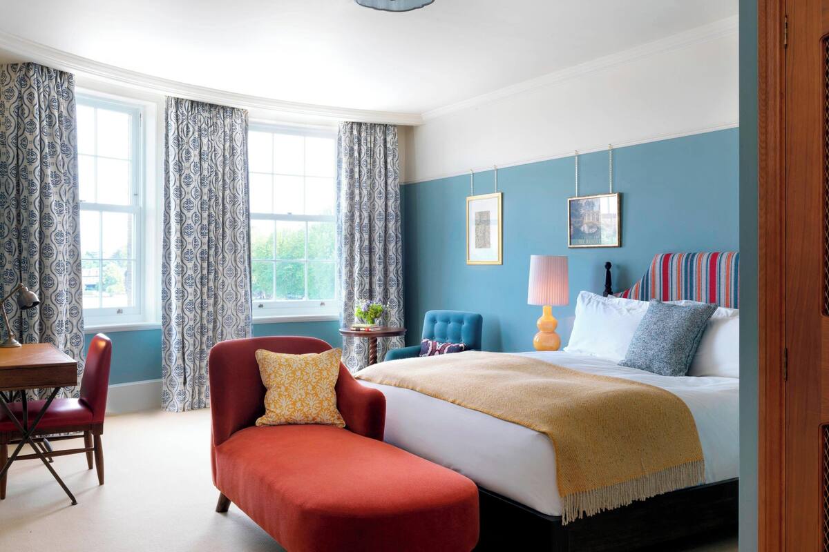 A bedroom with a bed, a red armchair, a small table with a lamp, and a window with curtains.