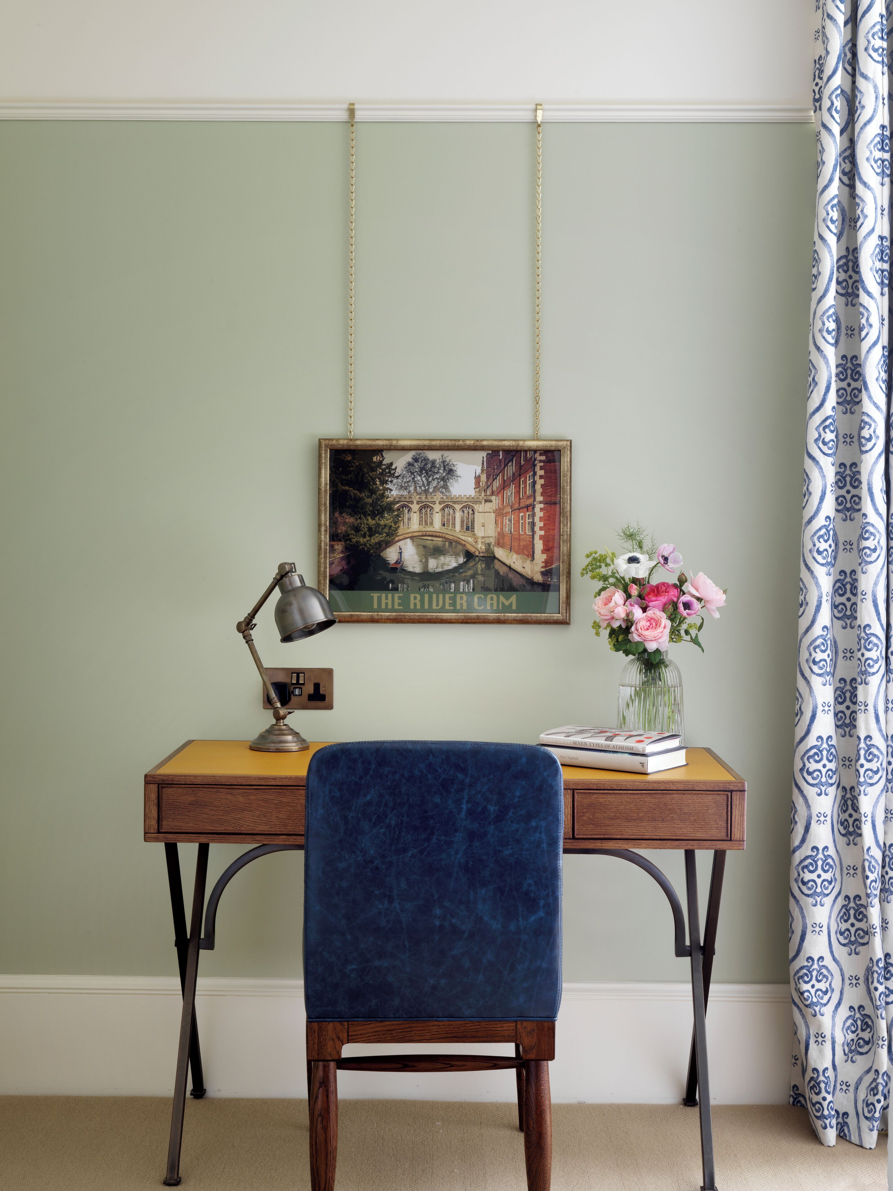 A desk with a blue chair, a framed picture, a lamp, and a vase with flowers.