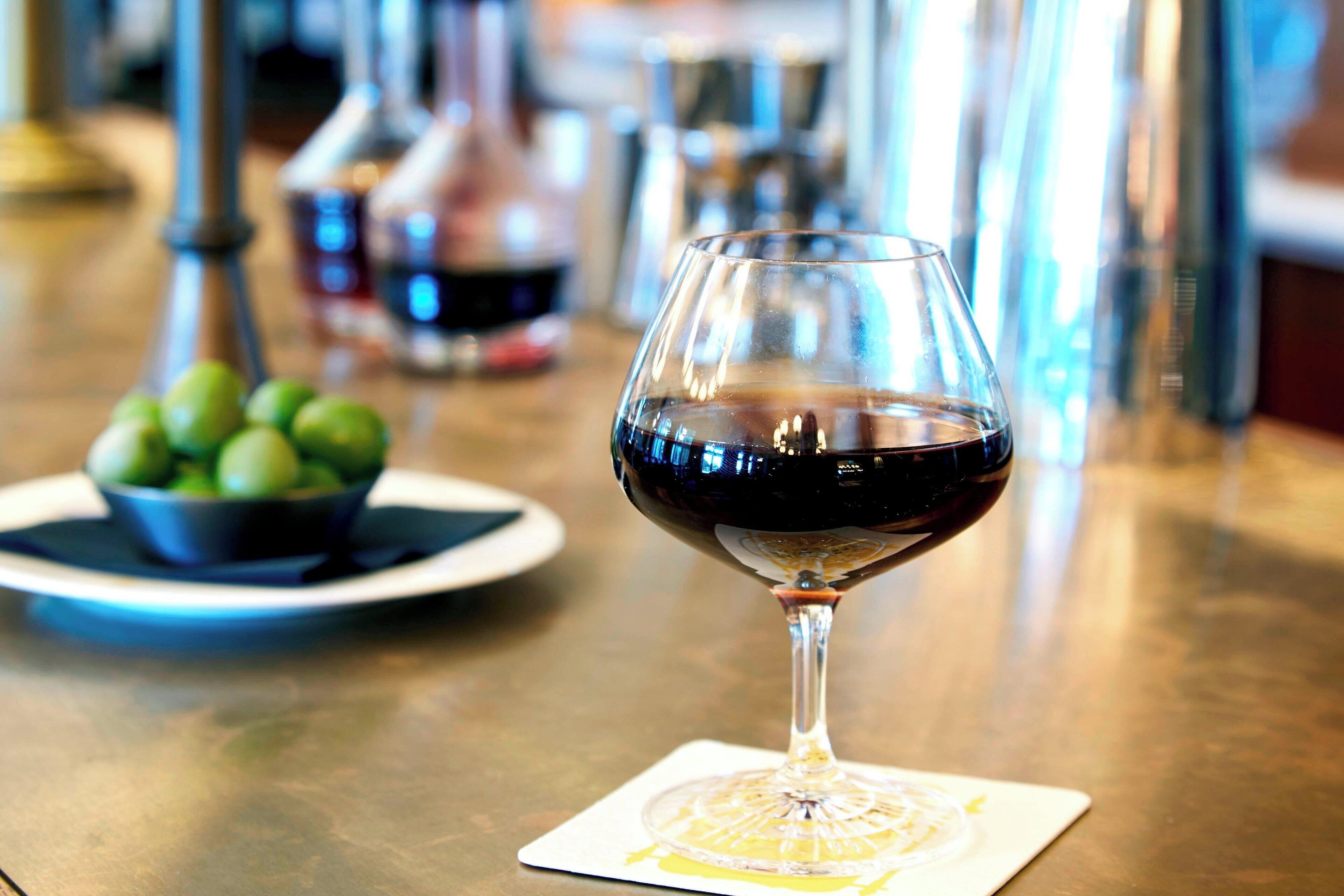 Um copo de vinho tinto sobre um apoio, com um prato de azeitonas verdes ao fundo.