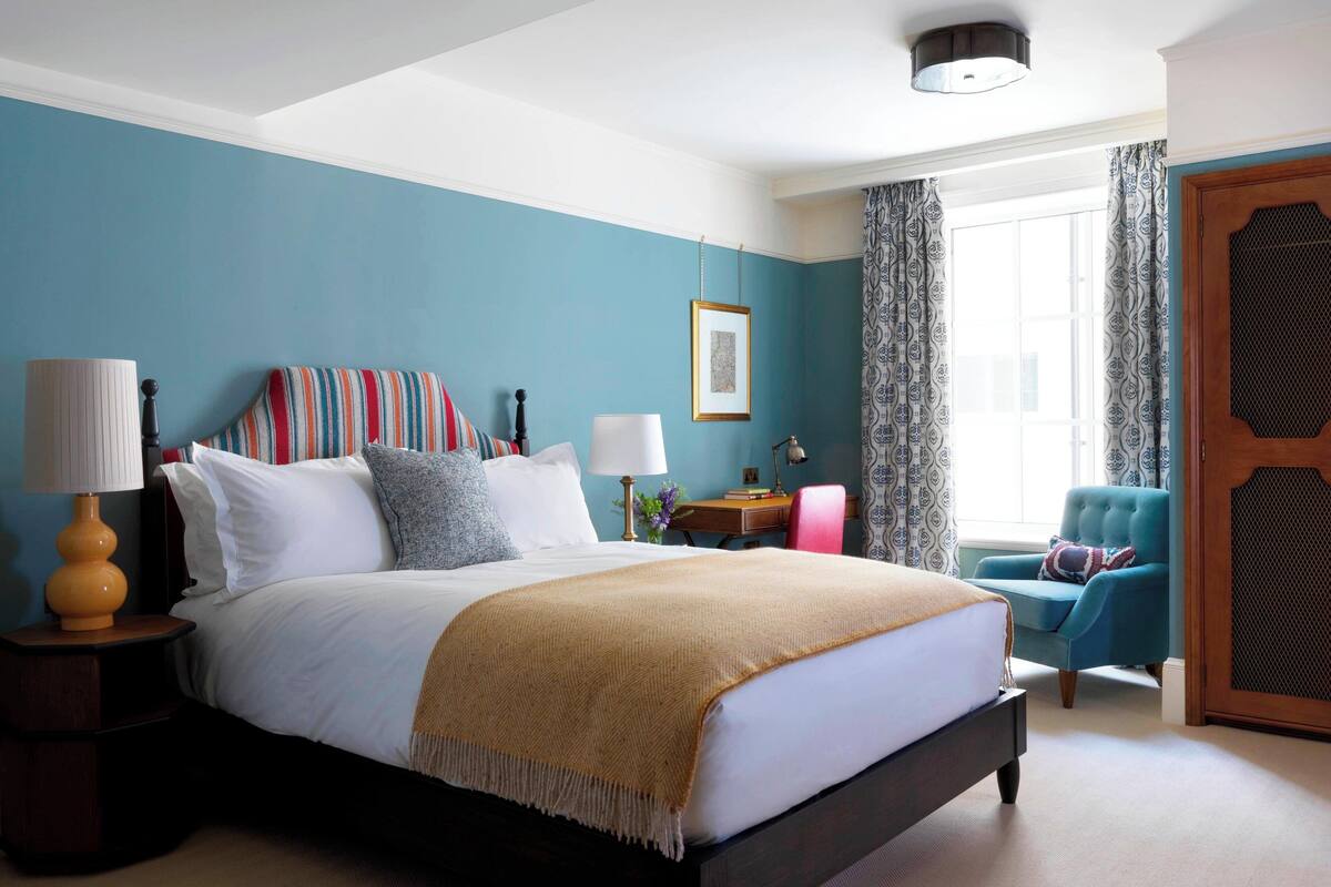 A neatly made bed with a white comforter and a beige throw blanket, a bedside table with a lamp, a chair with a patterned cushion, and a window with patterned curtains.
