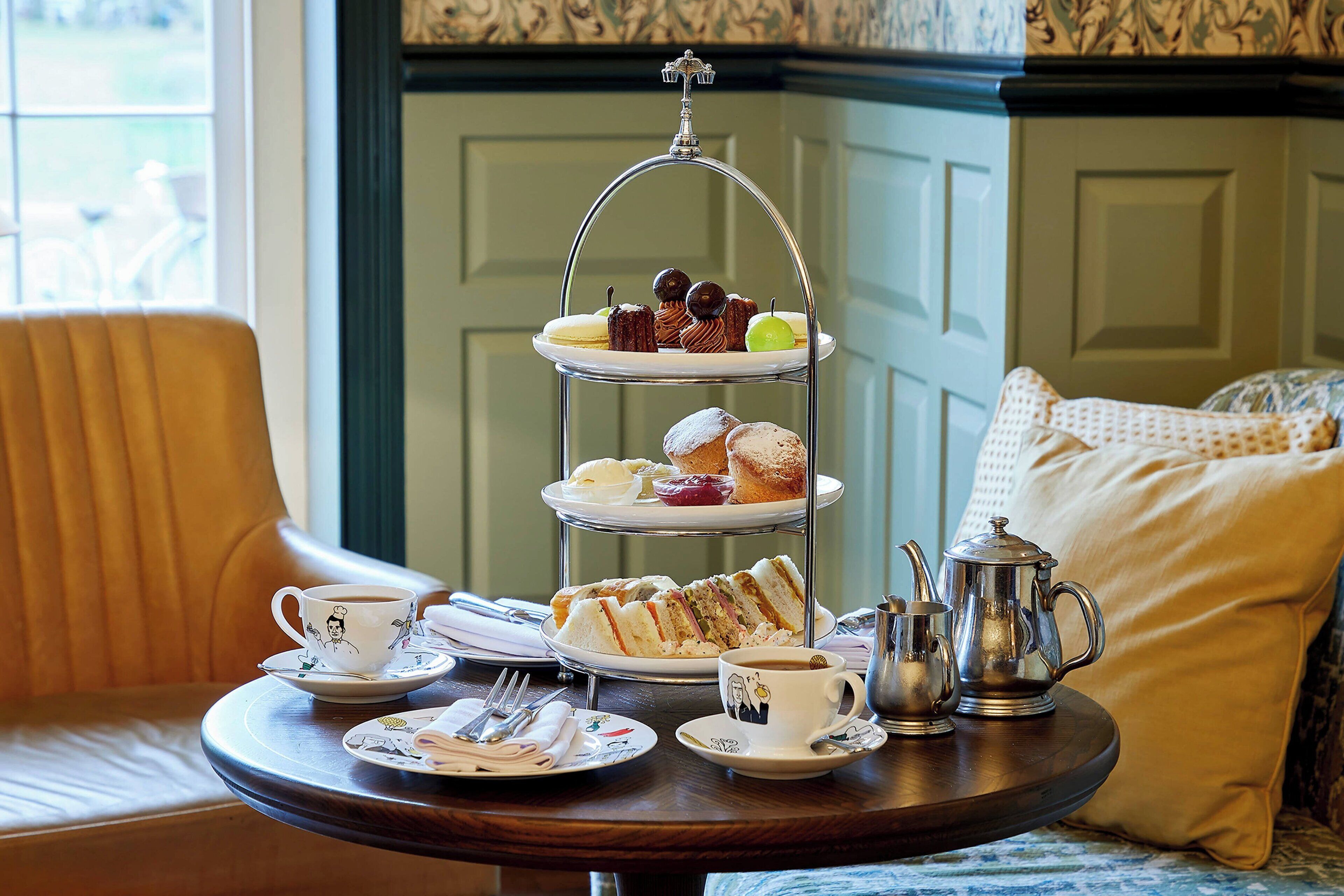 A three-tiered tea service tray with sandwiches and pastries, a teapot, cups, and cutlery on a table.