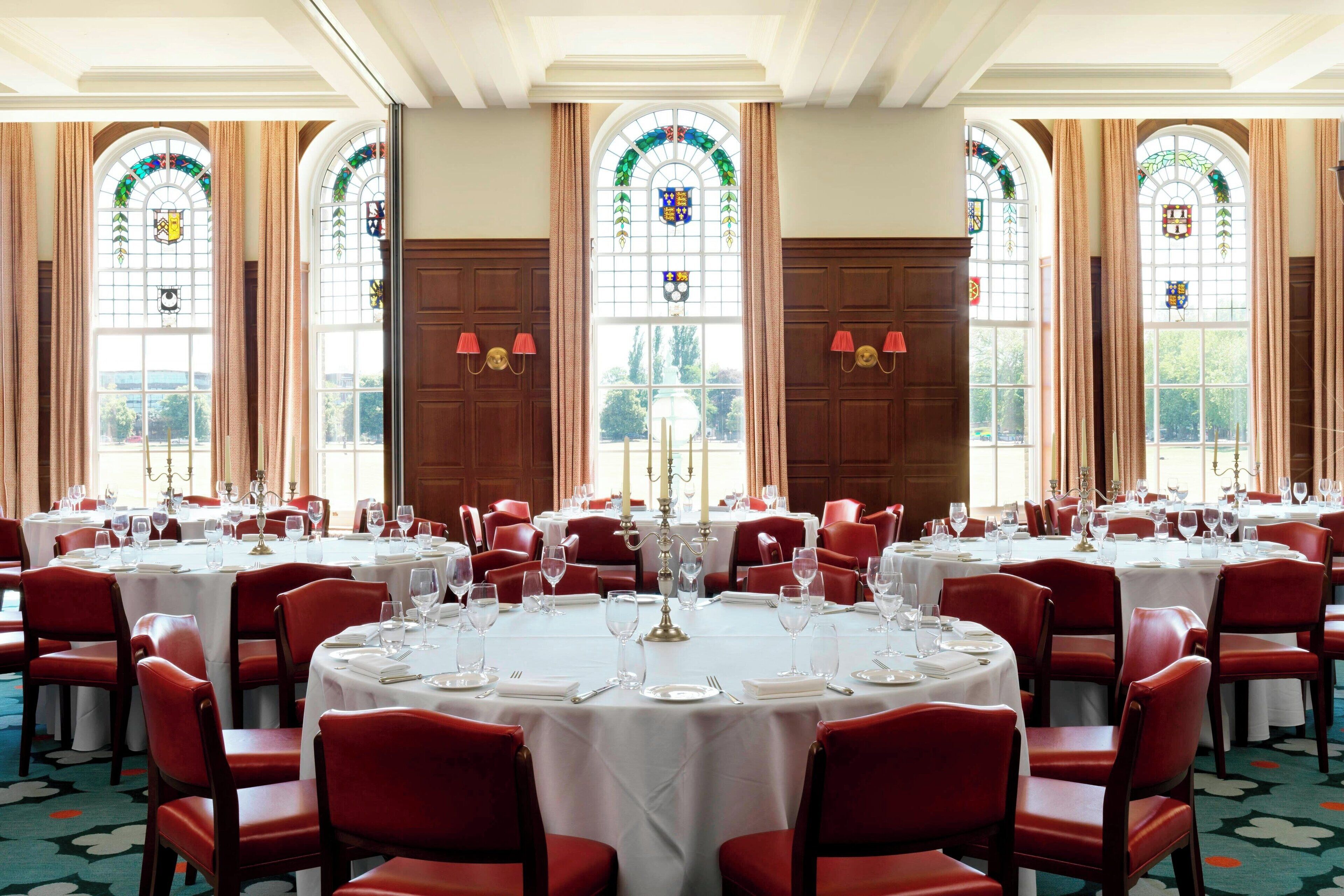 A formal dining room with round tables, red chairs, and large windows with stained glass.