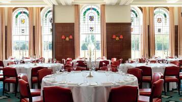 A formal dining room with round tables, red chairs, and large windows with stained glass.