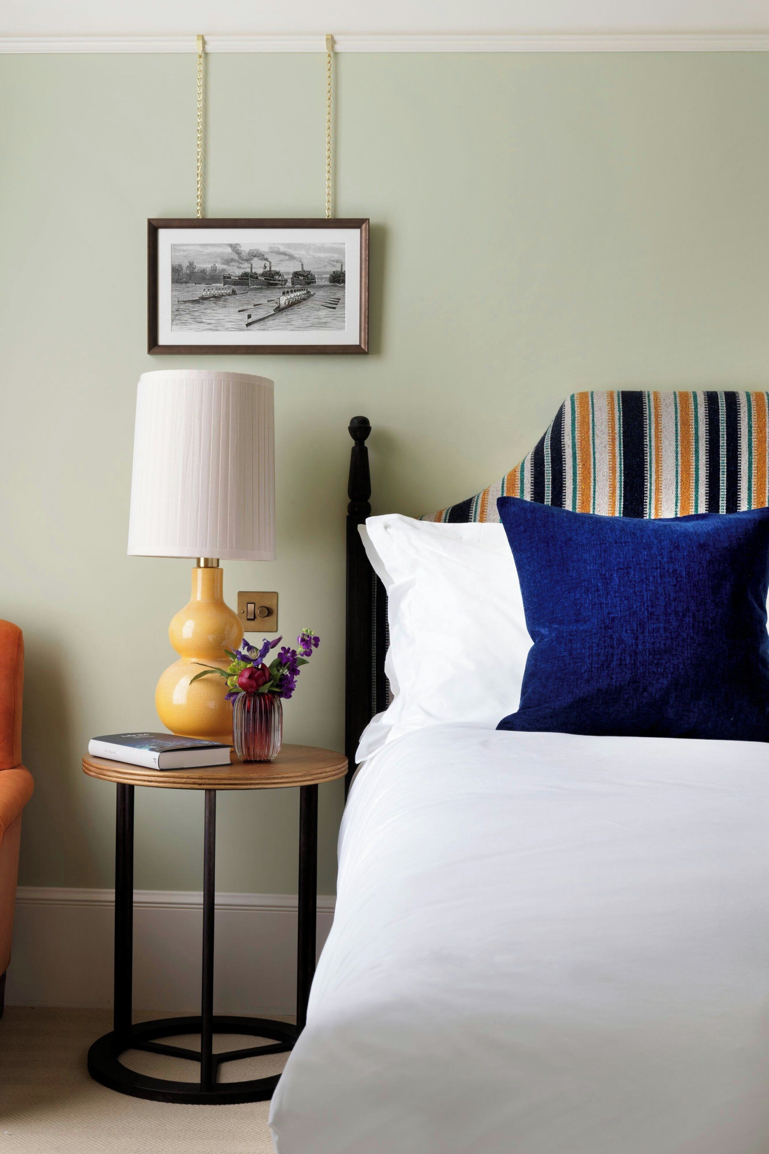 A neatly arranged hotel room with a bed, a bedside table with a lamp, a framed picture on the wall, and a striped pillow.