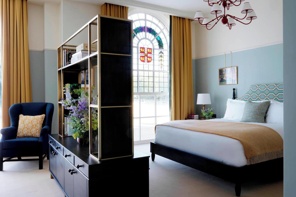 A bedroom with a bed, a chair, a bookshelf, a window with curtains, and a chandelier.