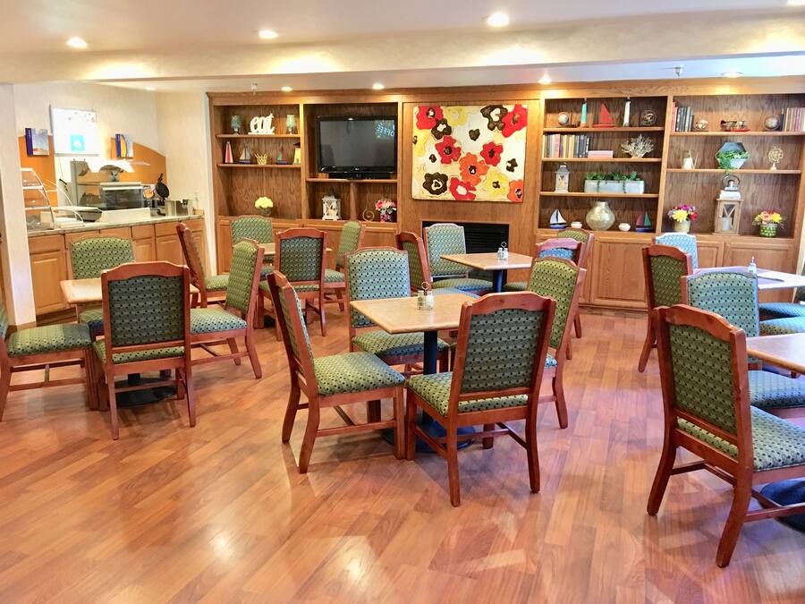 Un comedor con mesas y sillas de madera, un televisor, y estanterías con libros y objetos decorativos.