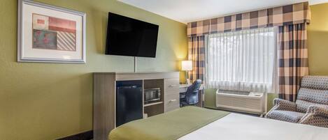 A hotel room with a bed, a desk with a computer, a flat-screen TV, a microwave, and a window with curtains.