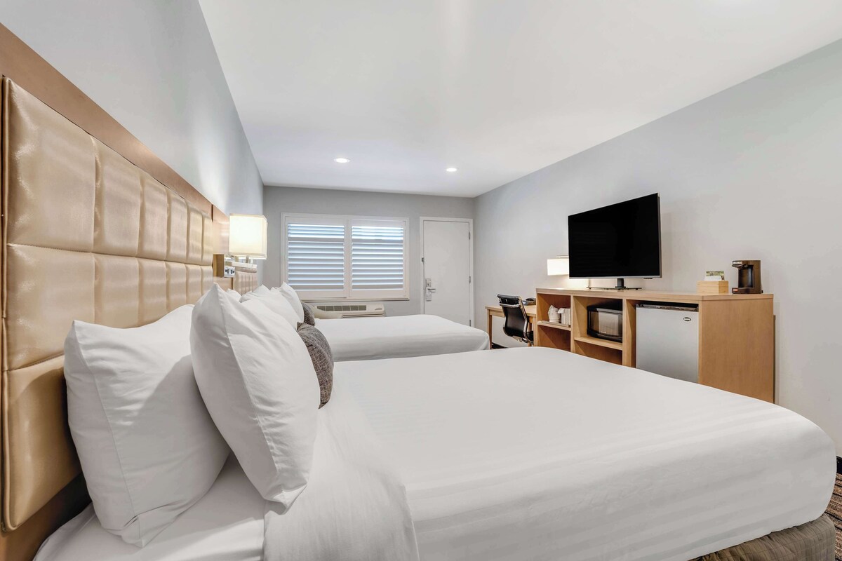 Ein Hotelzimmer mit zwei Betten, einem Fernseher, einem Schreibtisch mit Computer und einer kleinen Küchenzeile.