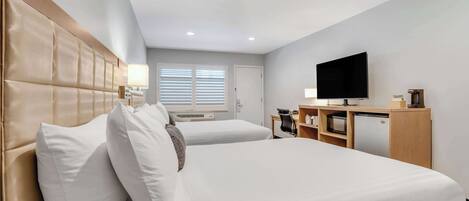 A hotel room with two beds, a TV, a desk with a computer, and a small kitchenette.