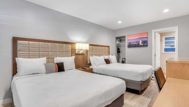 Habitación de hotel con dos camas, cabecera de madera, lámparas de noche, mesitas de noche, un escritorio y un cuadro de paisaje urbano en la pared.