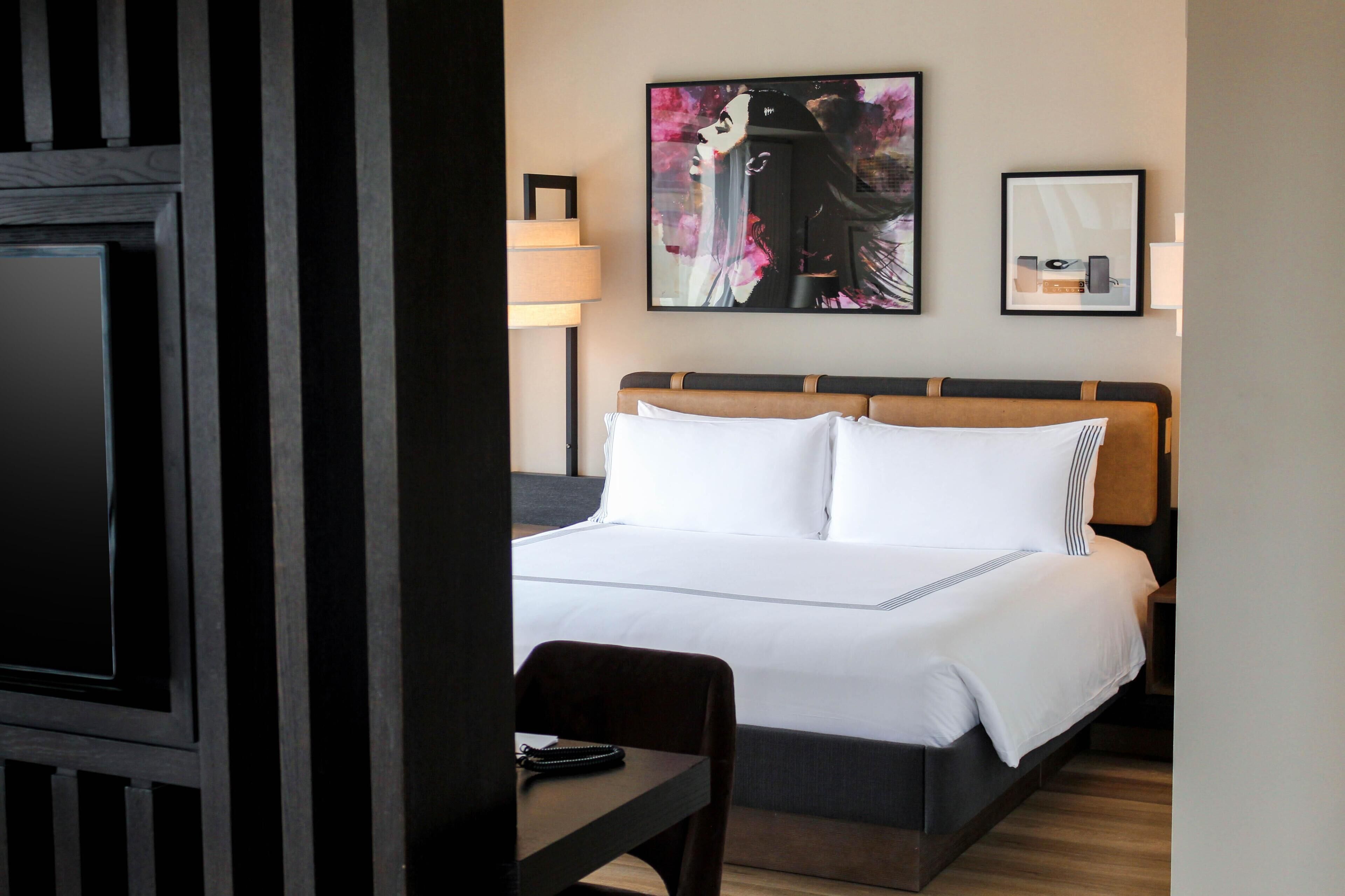Ein Hotelzimmer mit einem großen Bett, einem Schreibtisch und einem Stuhl. An der Wand hängt ein Gemälde und über dem Bett ein gerahmtes Bild.
