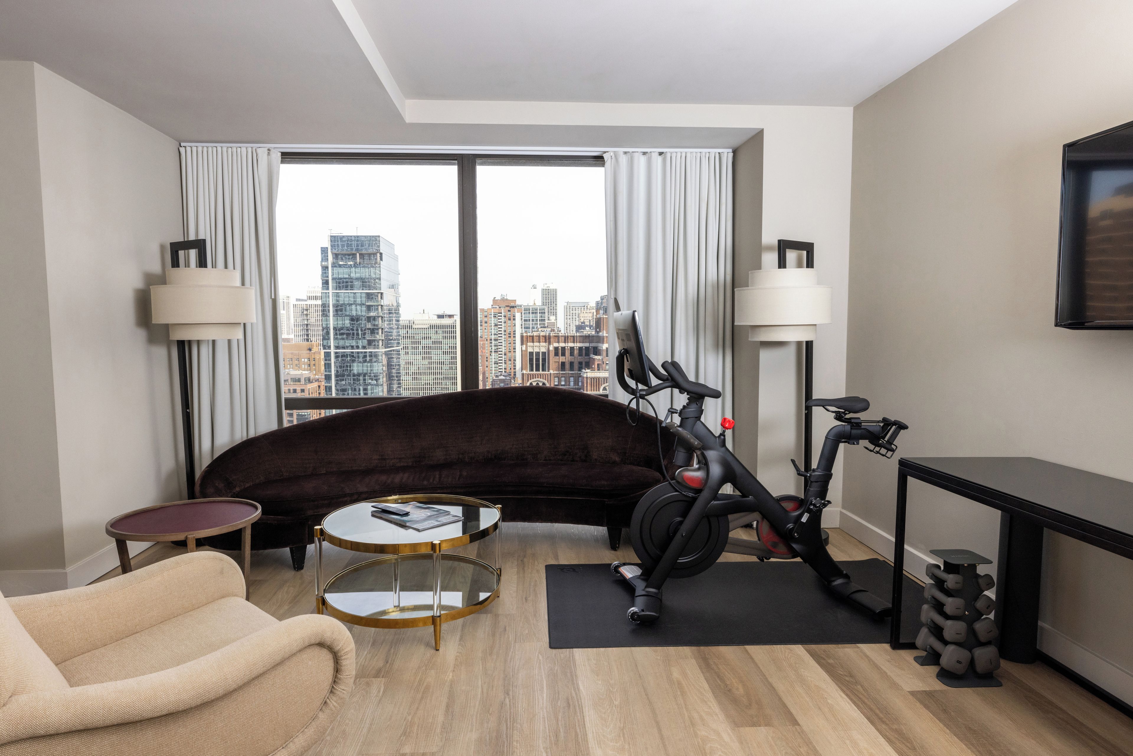 A modern hotel room with a large window, a sofa, a small round table, an exercise bike, and a TV mounted on the wall.