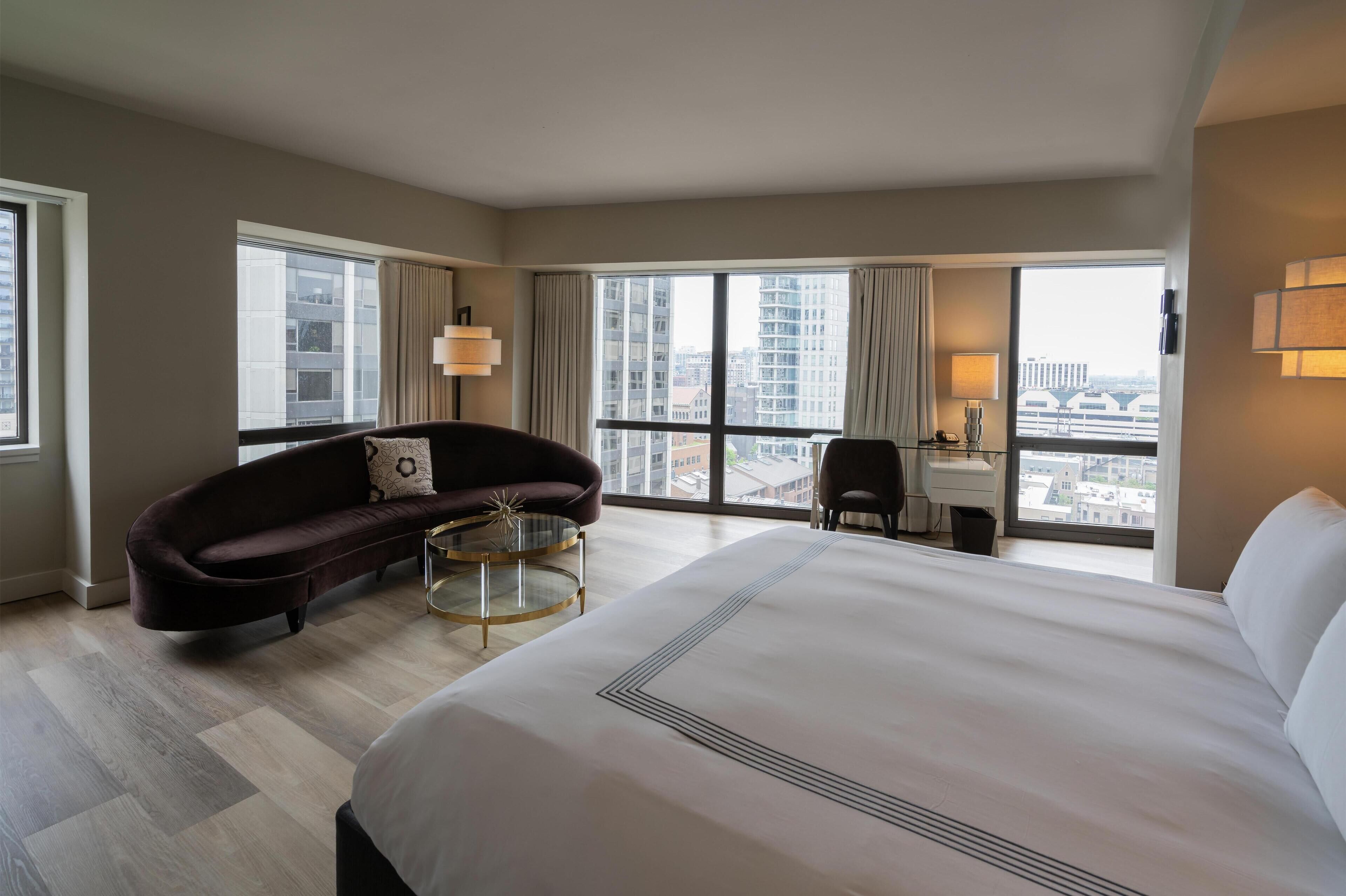 Habitación de hotel con una cama grande, un sofá oscuro, una mesita redonda pequeña, un escritorio con una silla y un amplio ventanal con vista a la ciudad.