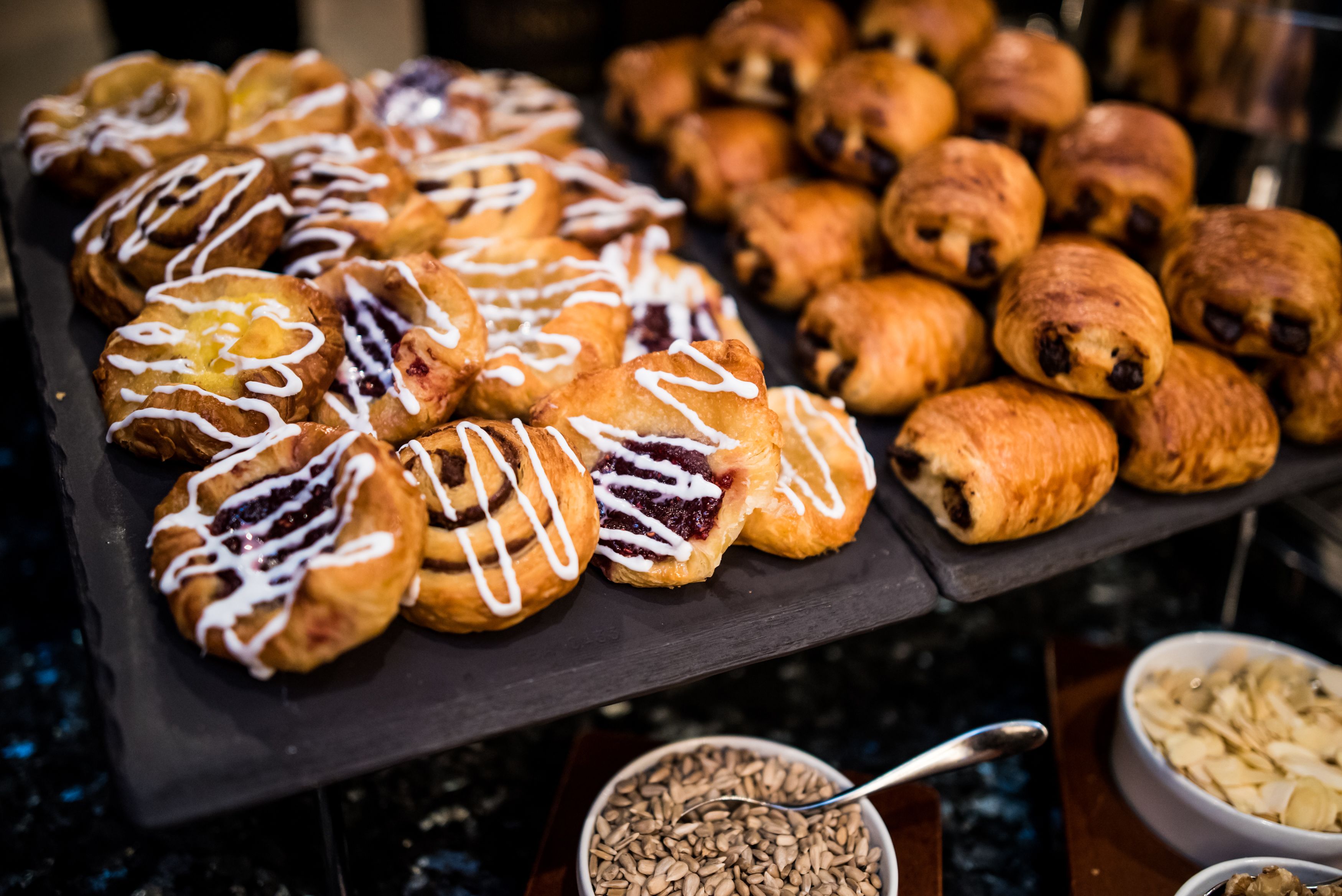 Eine Auswahl an Gebäck liegt zur Schau, darunter Croissants und Zimtschnecken, im Vordergrund steht eine Schüssel mit Granola.