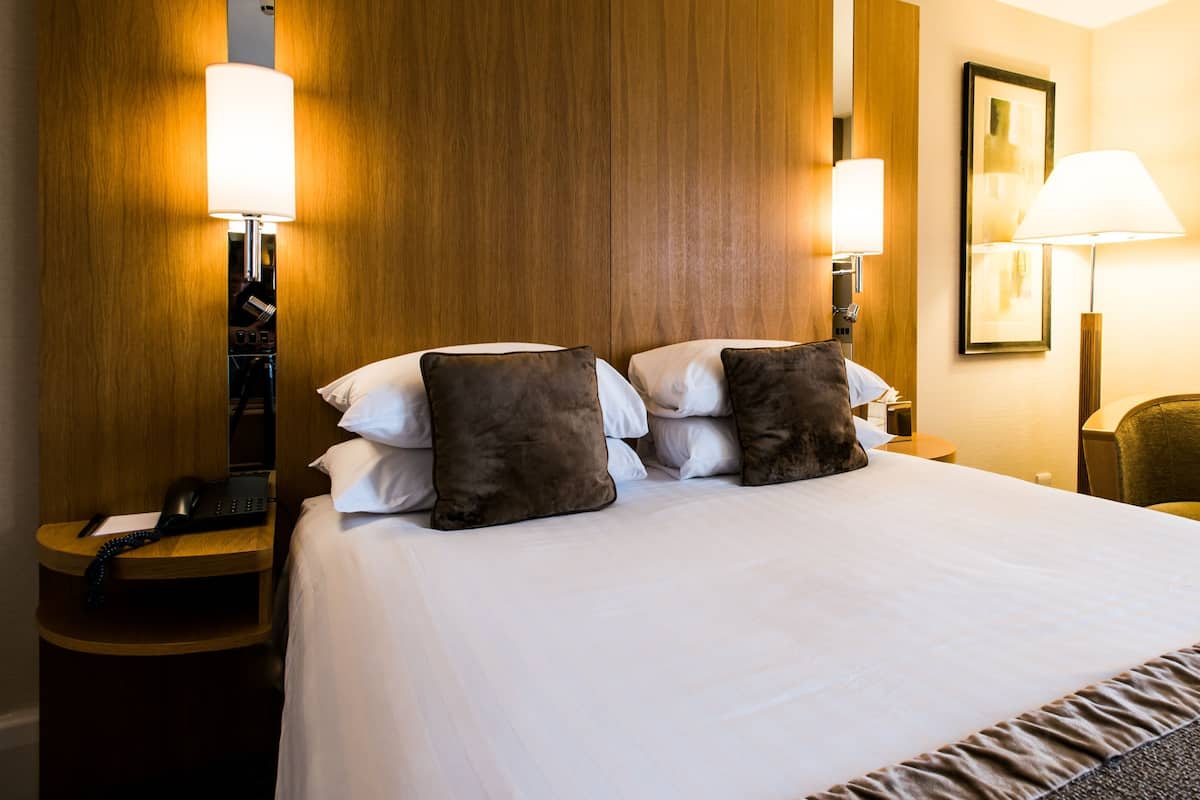 Ein Hotelzimmer mit einem Bett, zwei Nachttischlampen, einem Telefon, einem Sessel und einem gerahmten Bild an der Wand.