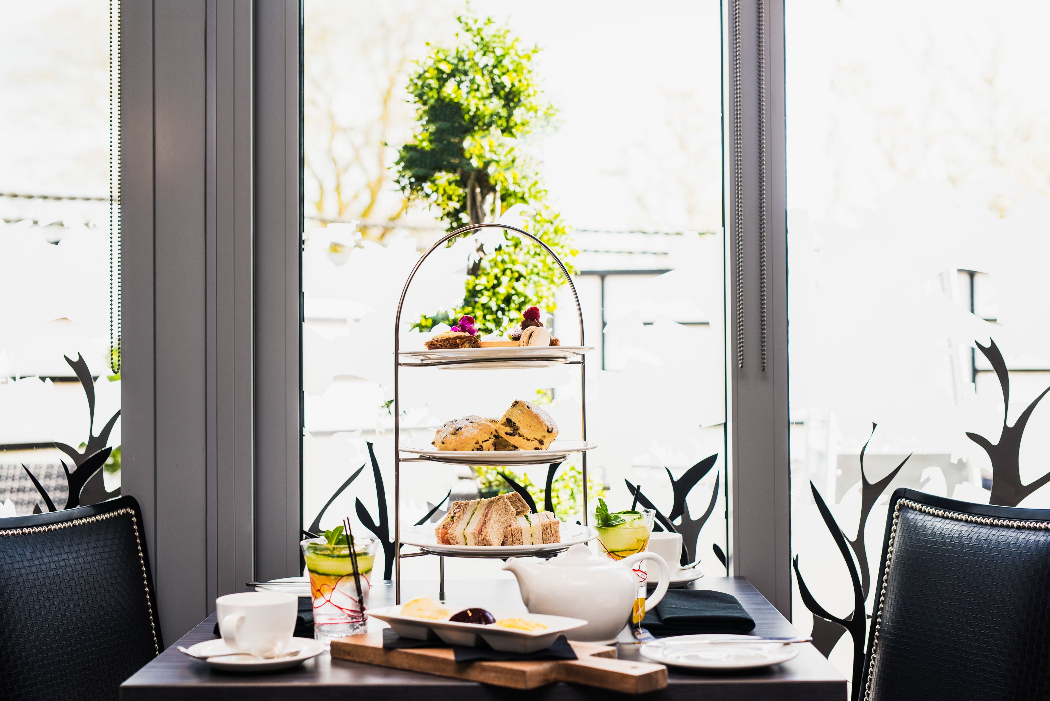 Un plateau à trois niveaux, garni de viennoiseries et de sandwichs, une théière et un verre de thé glacé, sont disposés sur une table près d’une fenêtre donnant sur un paysage verdoyant.