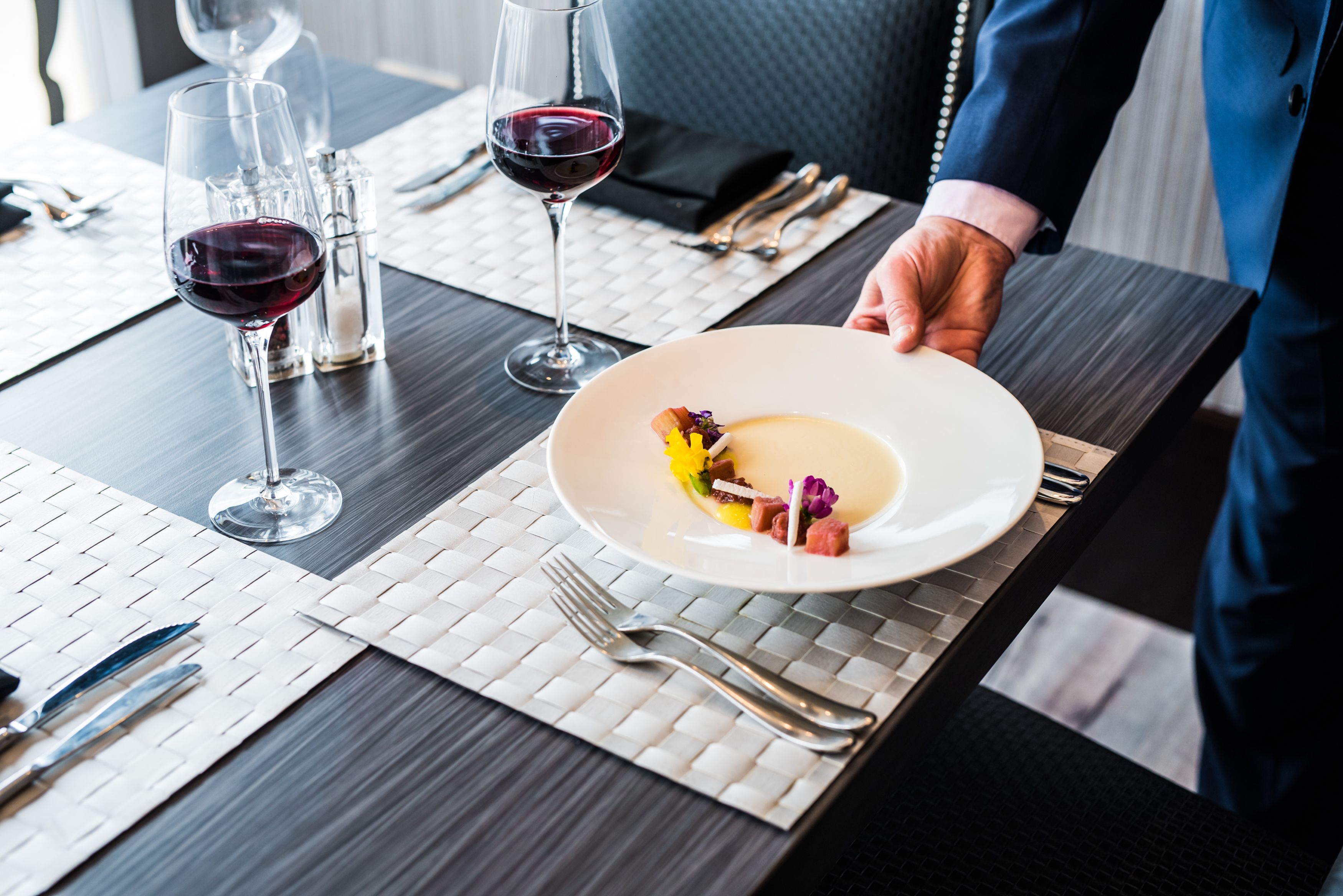 A dining table with a white placemat, a plate of food, and two glasses of red wine.