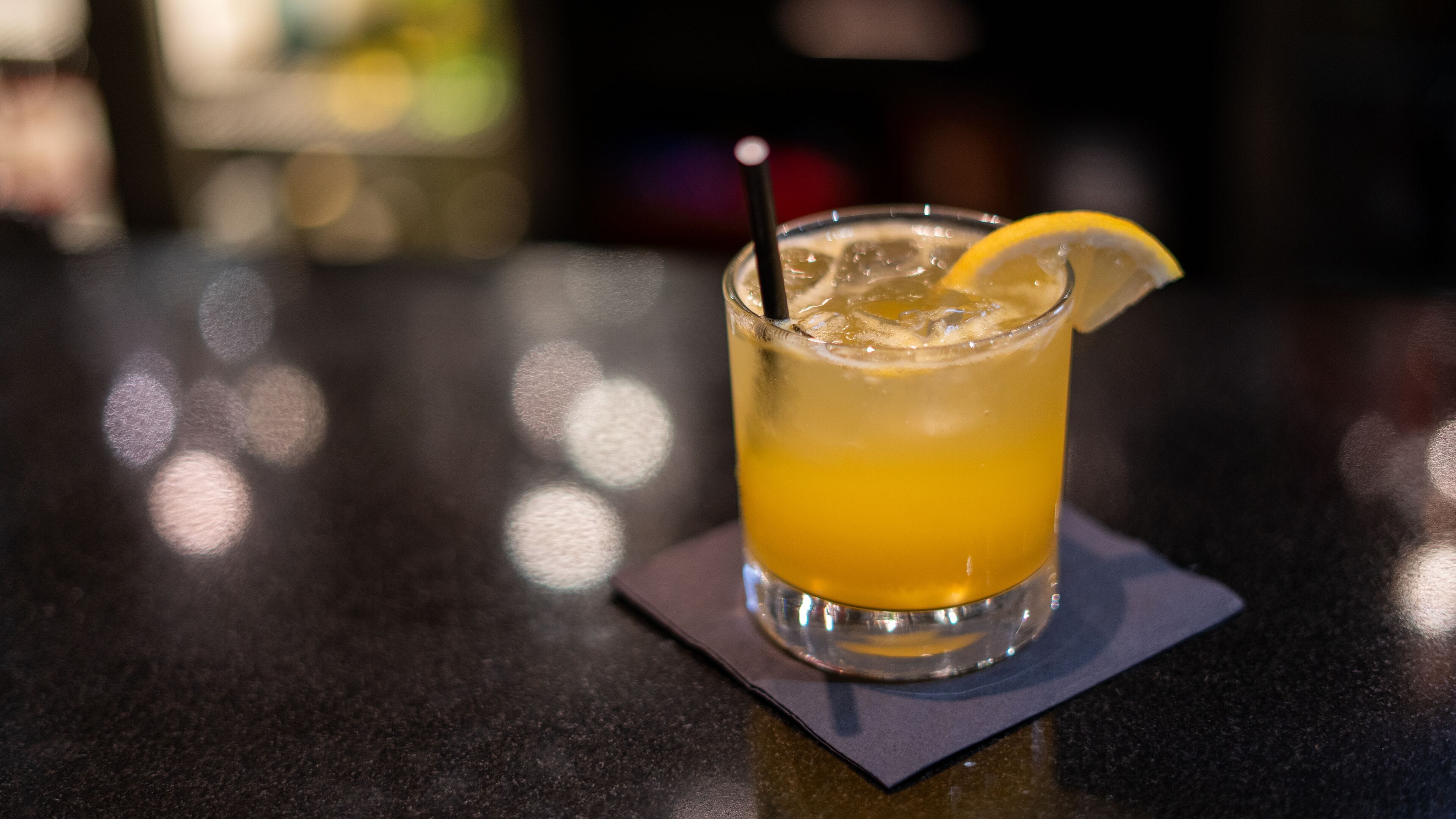 A glass of orange beverage with a lemon slice and a straw on a dark surface.