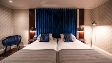 Una cama doble con una colcha estampada, dos mesitas de noche con lámparas, una silla azul y una cortina azul.