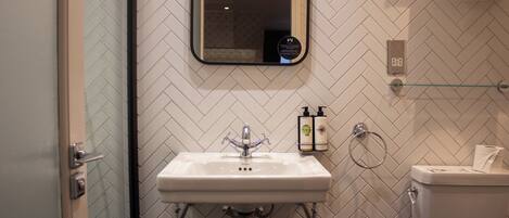 A bathroom with a white herringbone tile backsplash, a white sink with chrome fixtures, a glass shower enclosure, and a toilet with a towel rack above it.