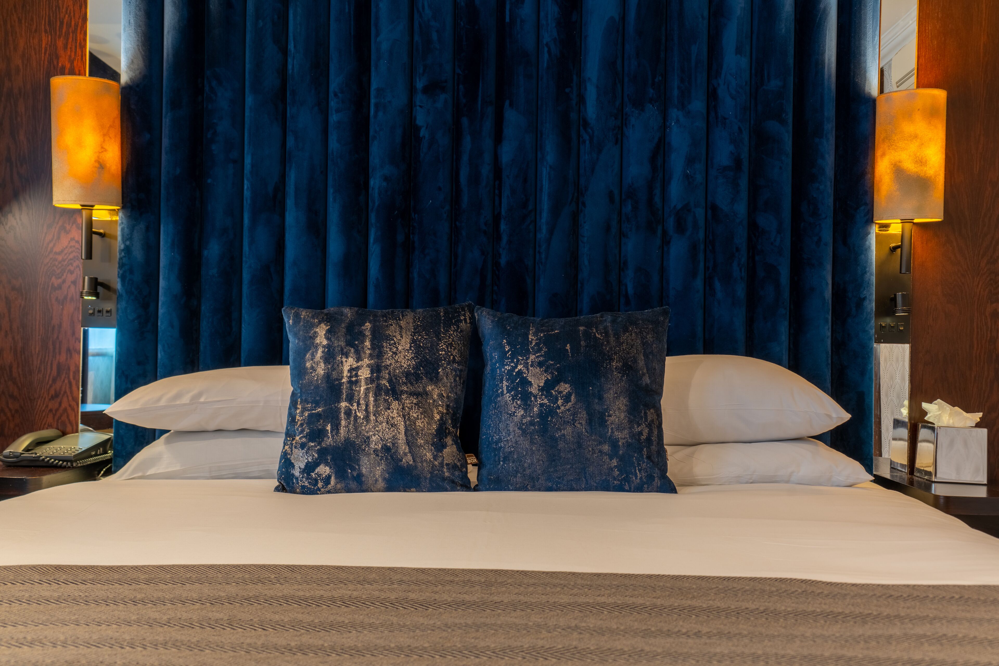 A hotel room with a large bed, dark blue curtains, two bedside lamps, and a wooden nightstand with a phone.