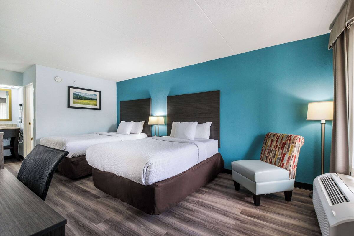 A hotel room with two beds, a desk, a chair, and a lamp.
