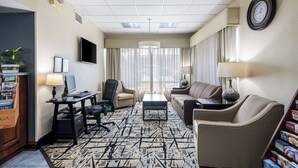 Ein Hotelzimmer mit großem Fenster, einem Flachbildfernseher, einem Schreibtisch mit Computer, einer Couch, einem Sessel und einem gemusterten Teppich.
