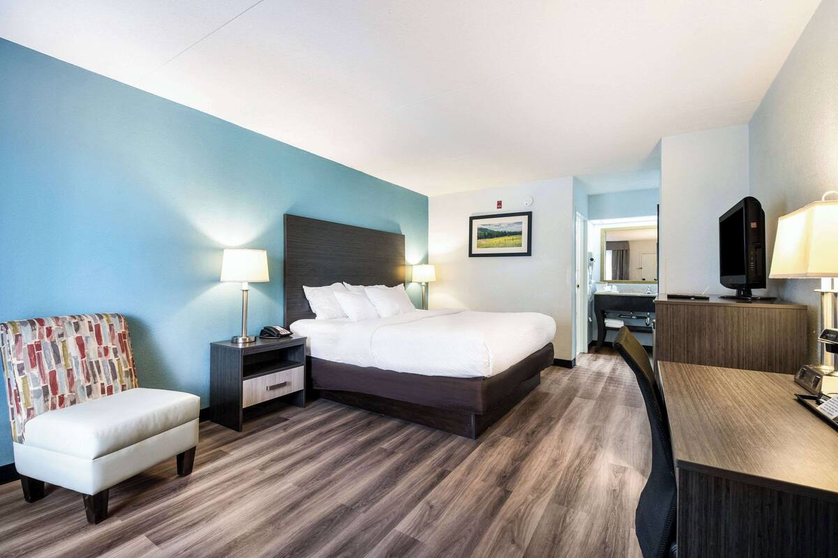 A hotel room with a bed, bedside table, lamp, chair, desk, and a television.
