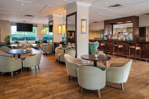 Una hall d'albergo con tavoli e sedie rotonde, un'area bar e ampie finestre che si affacciano sul verde.