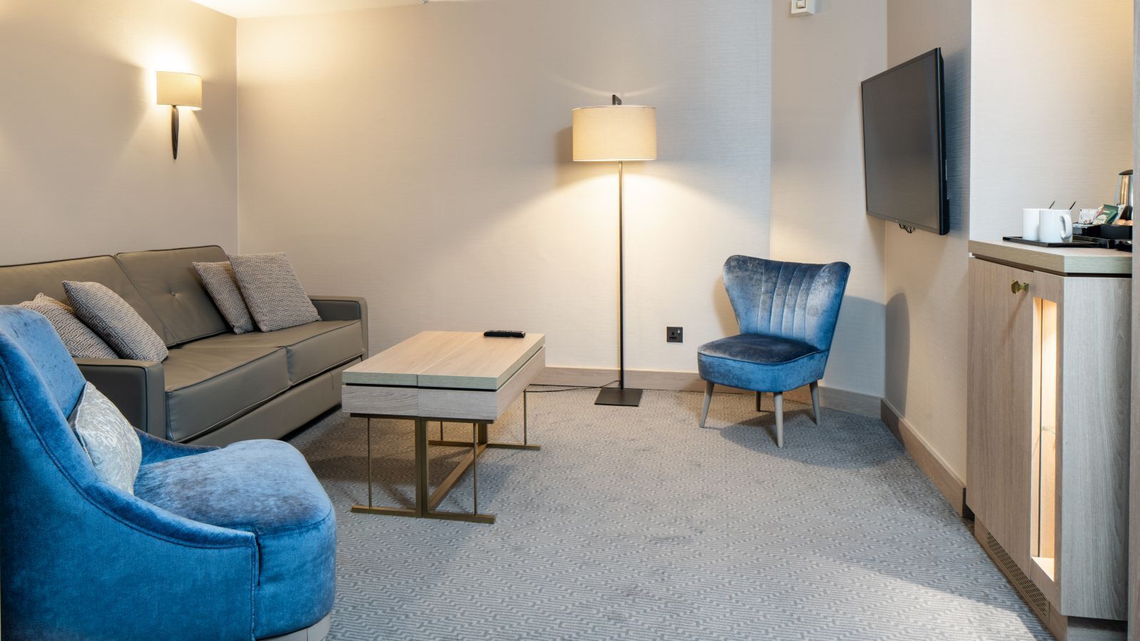 Een moderne woonkamer met een bank, twee fauteuils, een salontafel, een vloerlamp en een aan de muur bevestigde televisie.