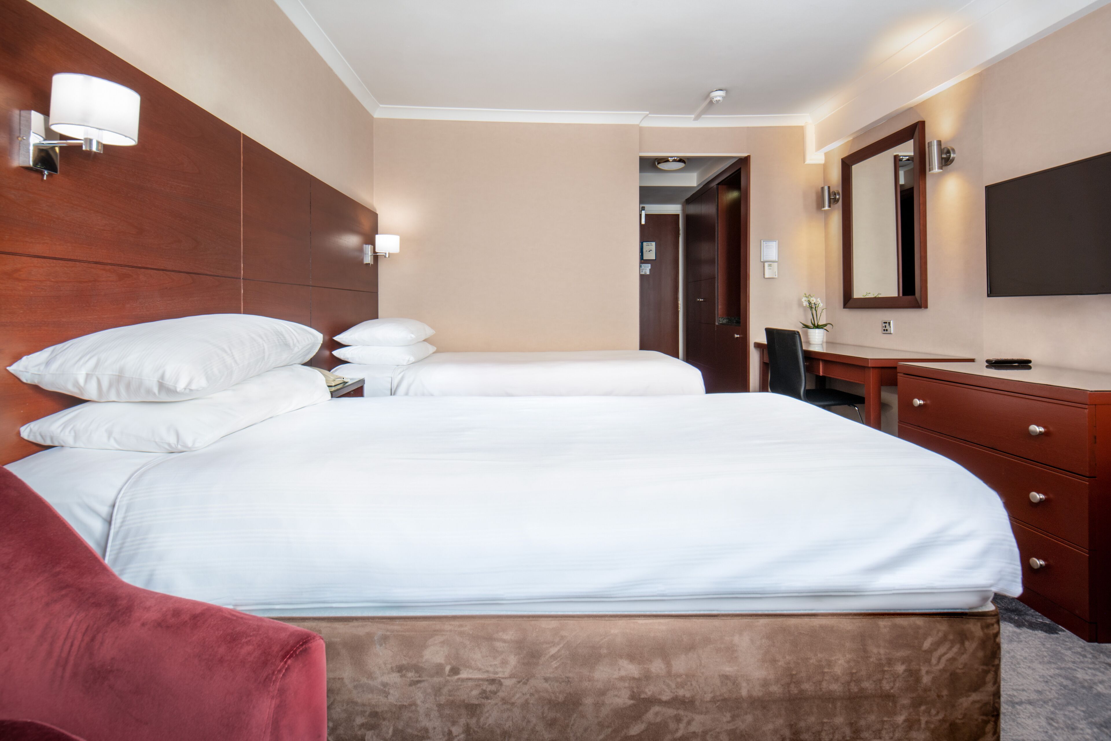 A hotel room with two beds, a wooden headboard, a desk with a chair, a dresser, and a flat-screen TV mounted on the wall.