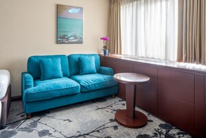 Ein smaragdgrünes Sofa mit zwei Kissen, ein kleiner runder Tisch, ein Bild von einer Strandansicht und ein Fenster mit beige farbenen Vorhängen.