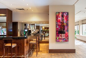 A modern bar area with wooden bar stools, a counter with various bottles, and a large abstract painting on the wall.