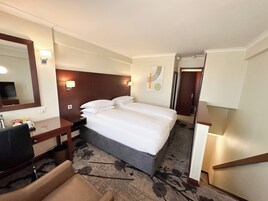 A hotel room with a large bed, a desk, a chair, a mirror, and a wall-mounted TV.