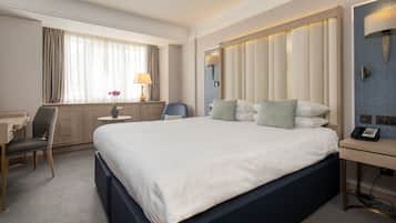A hotel room with a large bed, a desk with a telephone, a chair, a lamp, and a window with curtains.